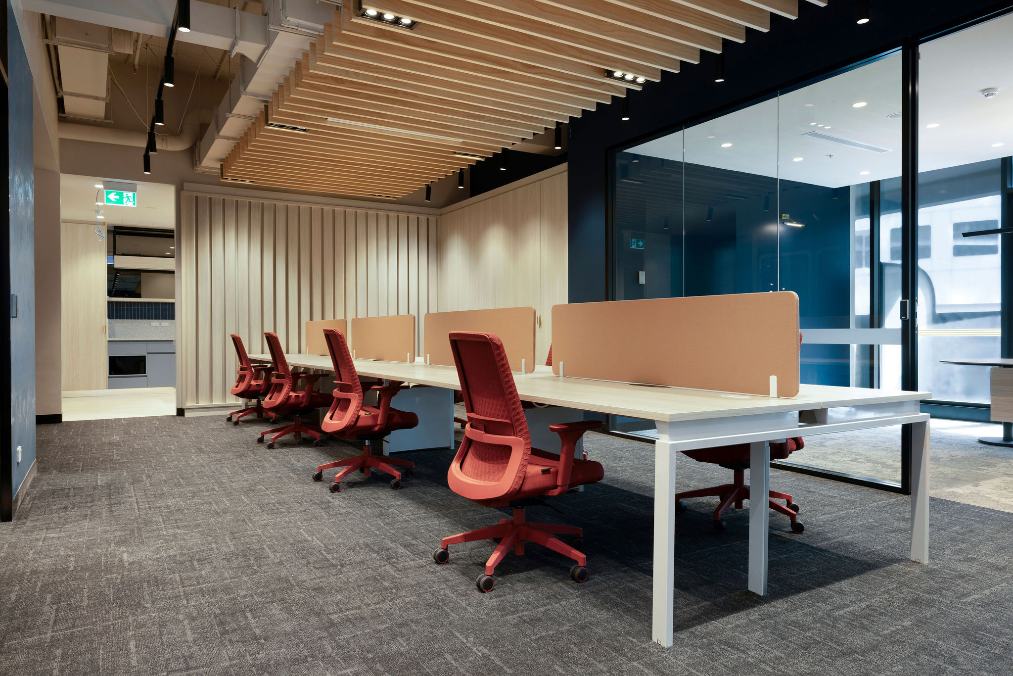 Open-plan office with white bench desks, red task chairs, acoustic desk screens and a timber batten ceiling